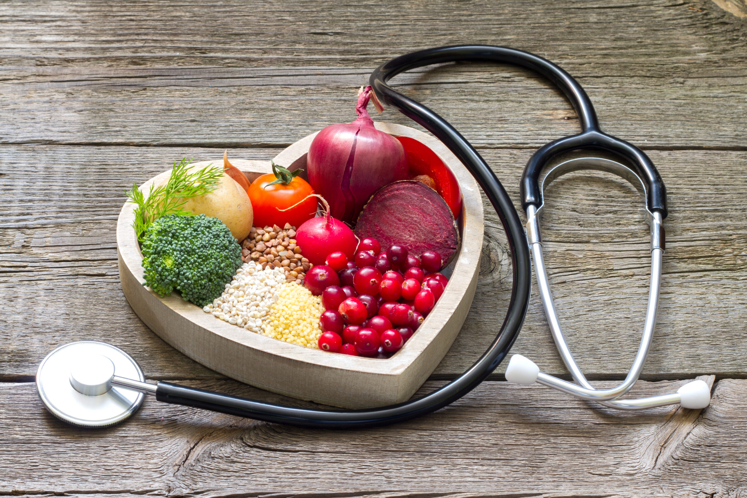 A heart shaped bowl full or healthy fruits and vegetables sits on a table next to a doctor's stethoscope.