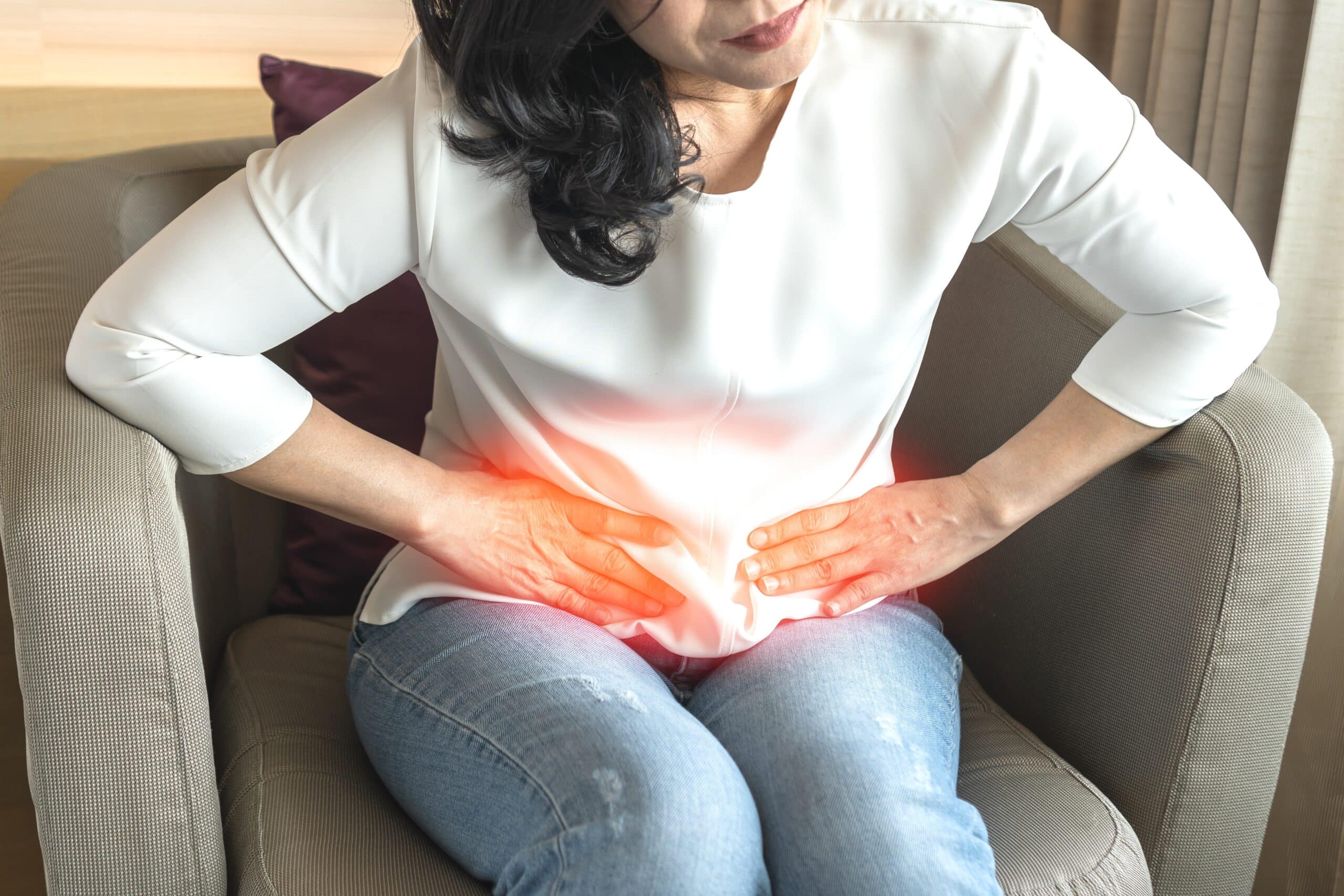 Woman in white blouse and blue jeans slouches over in pain suffered in the abdominal region.