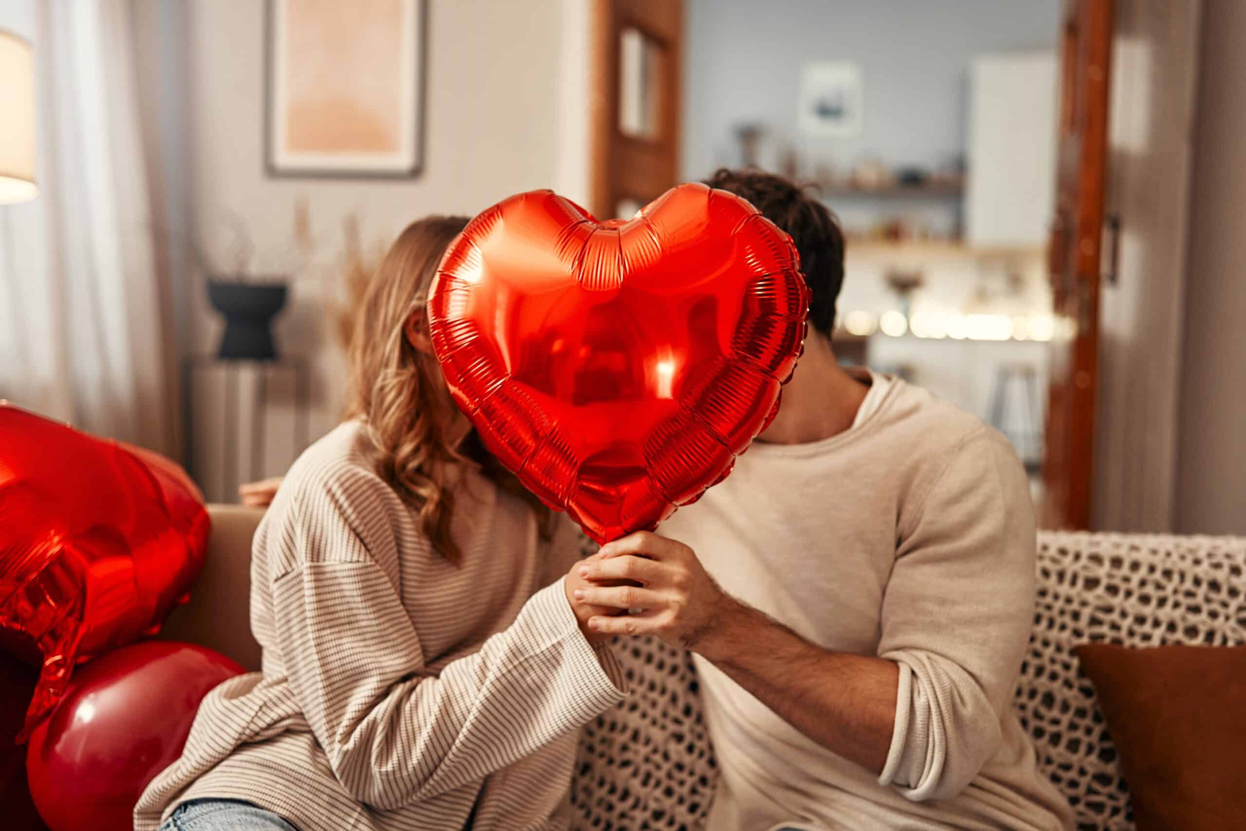 Young couple in love holding a heart-shaped balloon, covering themselves with it while kissing, sitting on the sofa in the living room at home