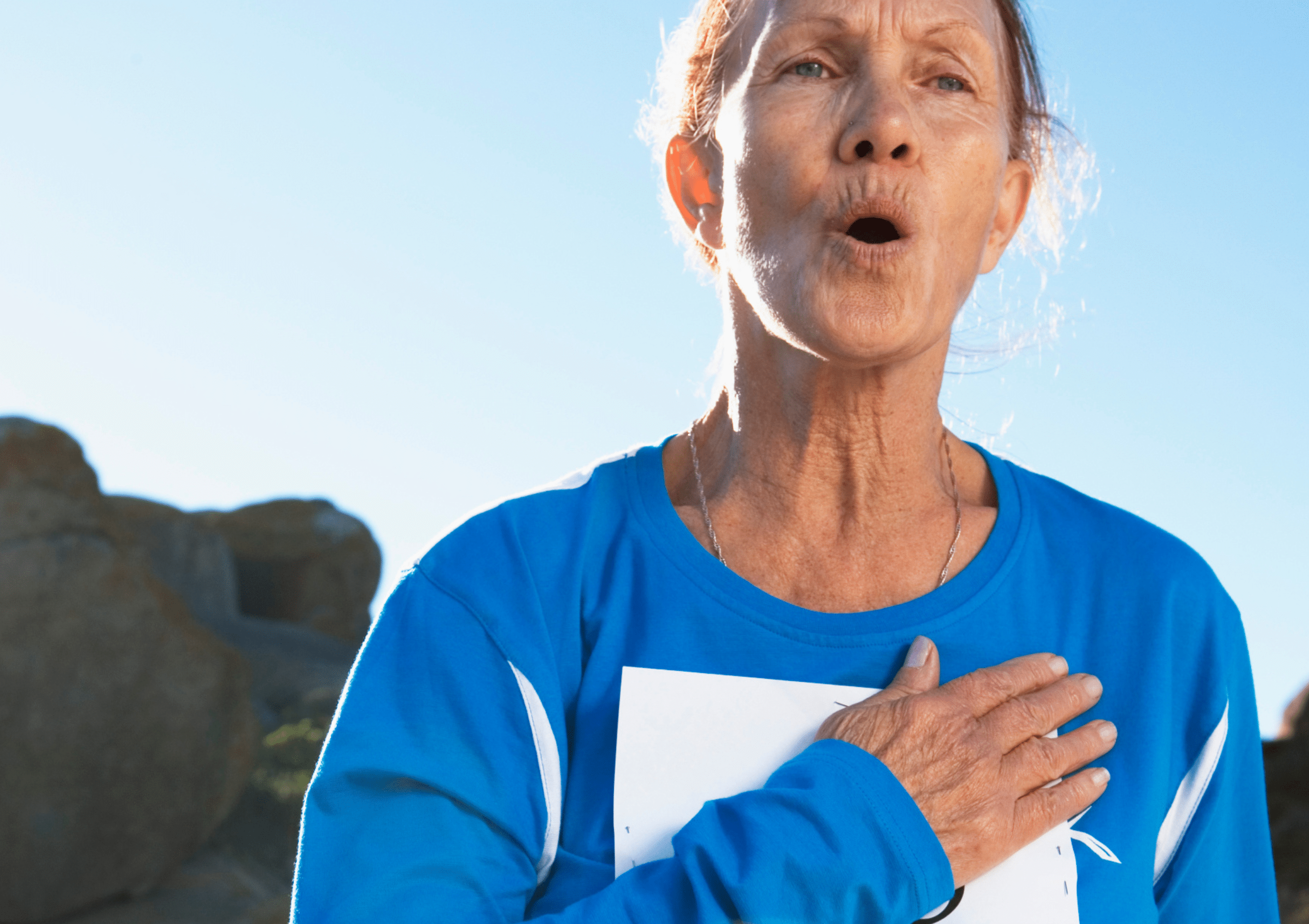 An older woman in blue marathon running gear holds her hand over her chest from shortness of breath.