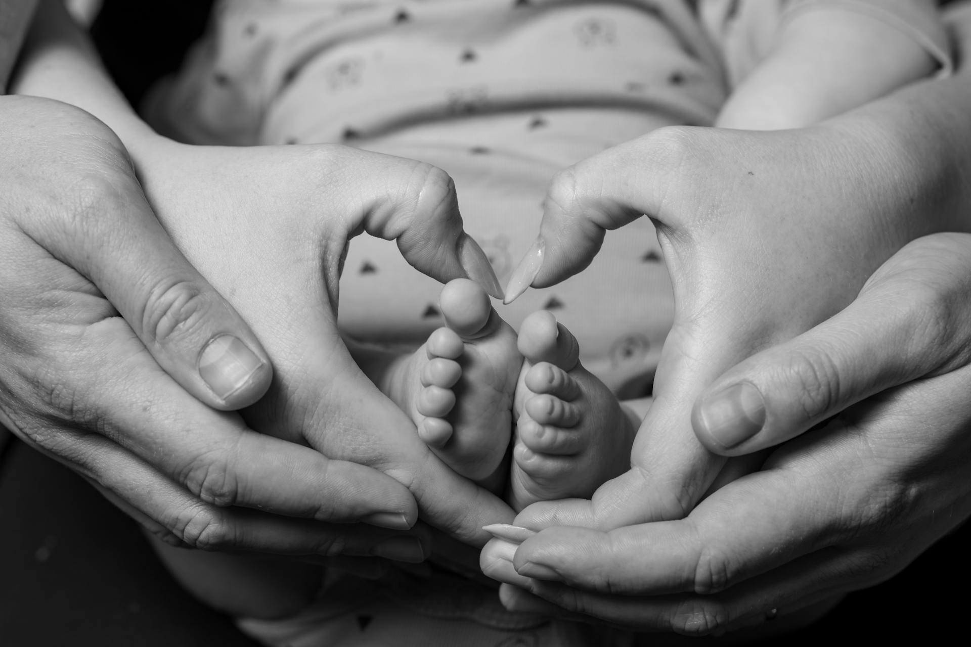 Black and white close-up of father and mother's hands making a heart shape around their infant's feet.