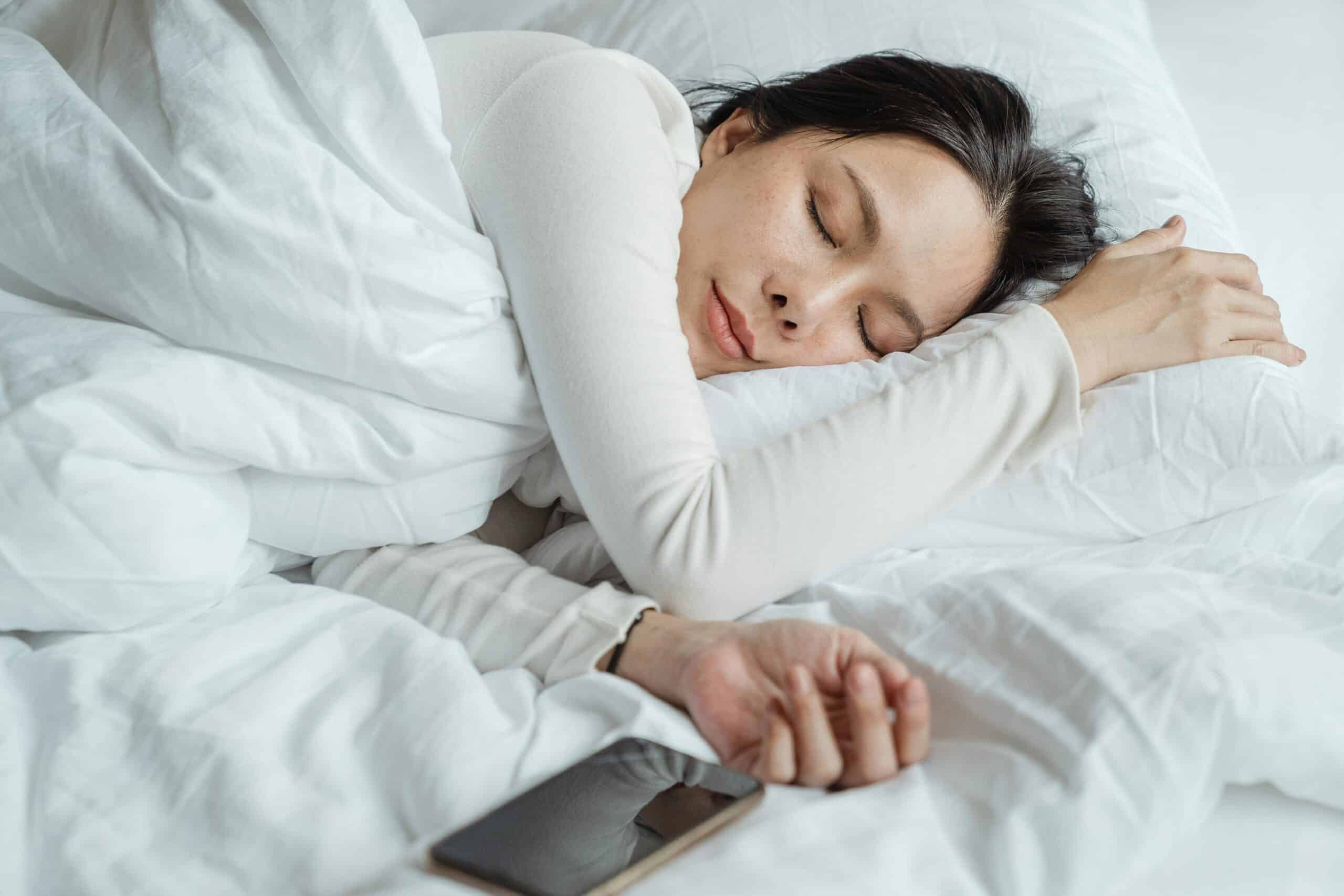 A woman with dark hair in white pajamas sleeps soundly and healthily on her side next to her cellular phone.