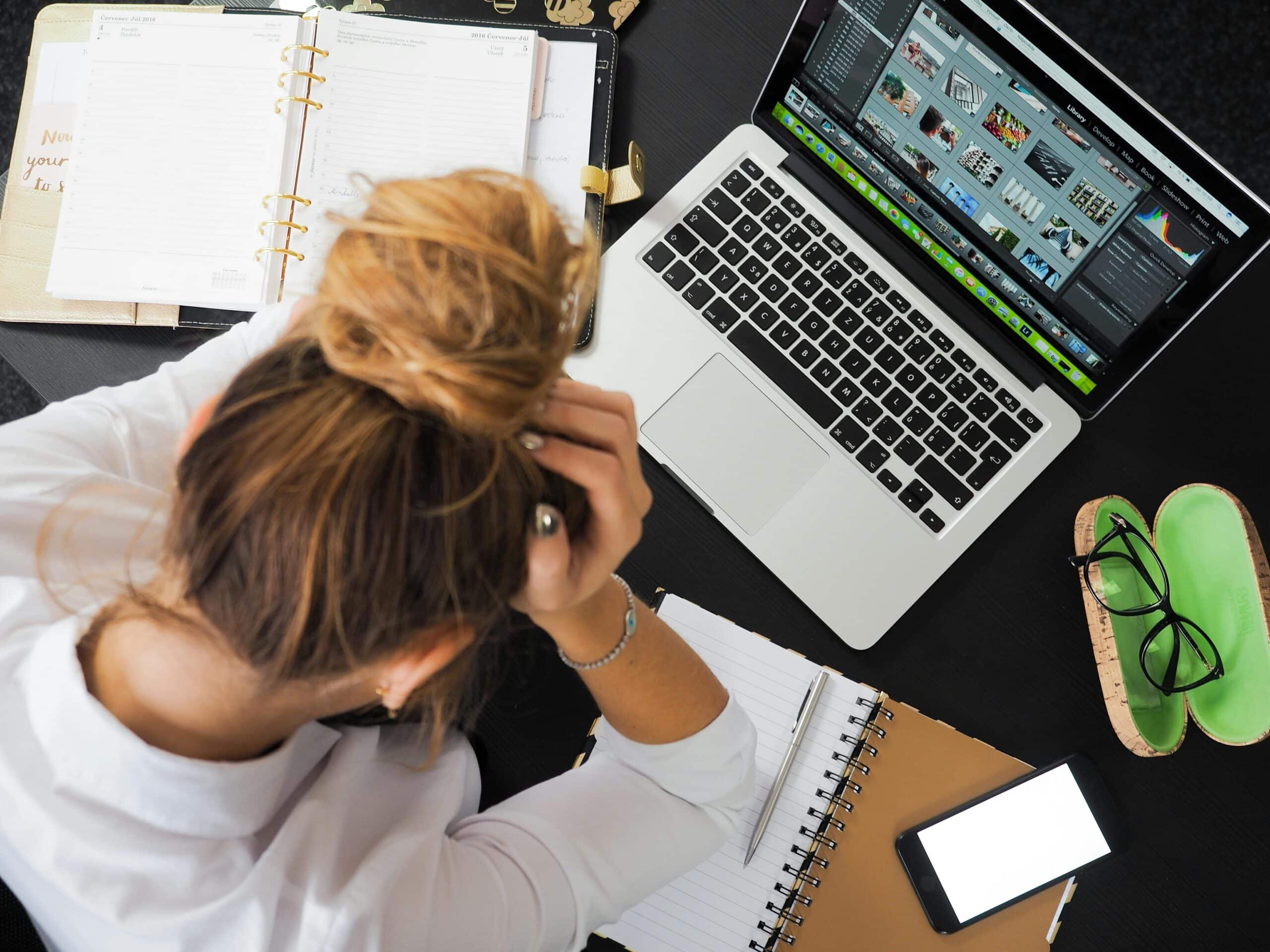 An over-the-shoulder view of a woman at a desk surrounded by her planner, computer and phone - burying her head in her hands from stress.