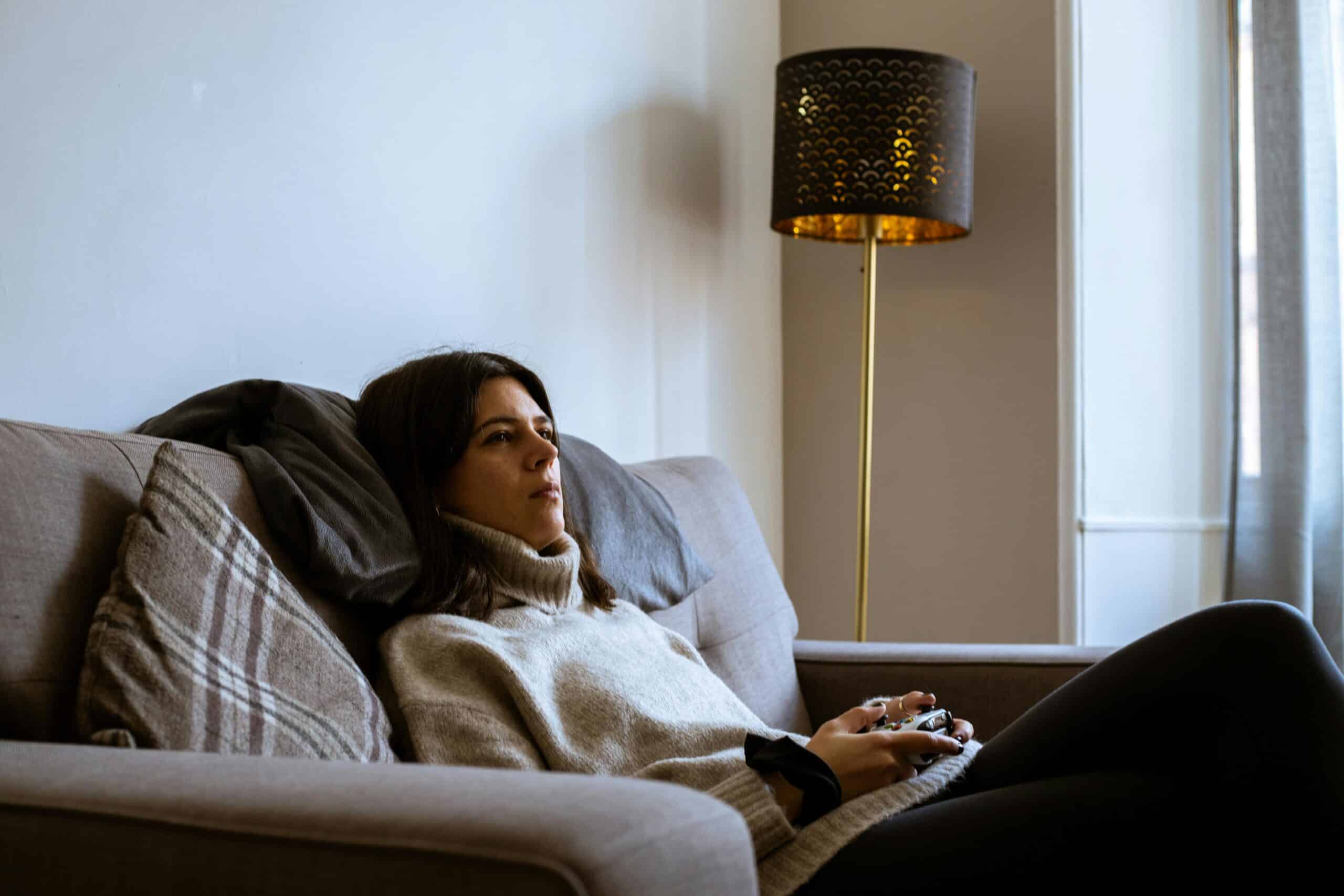 A woman in a comfy turtleneck leans back on the couch while holding a video game controller.